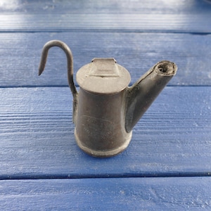 May include: A small, antique metal watering can with a curved handle and a spout. The can is a muted silver color, showing signs of age and wear. The lid is hinged, and the spout is slightly angled. The watering can is likely used for miniature plants or decorative purposes.
