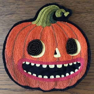 May include: An embroidered pumpkin patch featuring orange thread for the body, a green stem, and black beaded eyes. The pumpkin has a wide, grinning mouth with white teeth outlined in pink thread, all set against a black fabric backing.