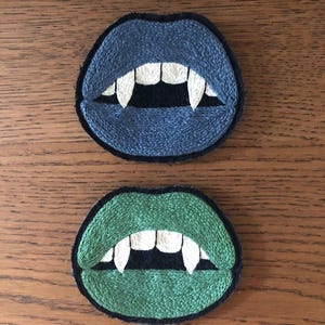 May include: Two embroidered patches shaped like open mouths with sharp white fangs. The top patch features blue lips, while the bottom patch features green lips. Both designs include black detailing around the teeth and a dark border outline.