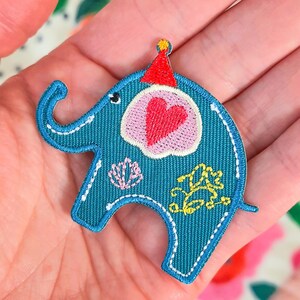 Könnte beinhalten: Ein blauer gestickter Elefanten-Patch mit einem rosa Herzen, gelben Blumen und einer rot-gelben Partymütze.