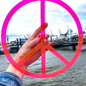 Könnte beinhalten: Ein leuchtend pinkfarbenes Friedenszeichen-Ornament mit orangefarbenem Zentrum, gehalten vor einem bewölkten Himmel und Wasser. Das Friedenssymbol besteht aus einem durchscheinenden Material und hat oben ein kleines Loch zum Aufhängen.