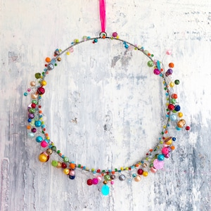 May include: A silver wire wreath decorated with colorful beads and sequins. The wreath is hanging from a pink ribbon against a white textured background.