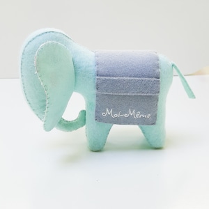 Può includere: Un giocattolo a forma di elefante in feltro azzurro con una sella grigia. L'elefante ha grandi orecchie e una piccola proboscide. La sella è ricamata con la scritta "Moi-Même". Il giocattolo è in morbido feltro e ha cuciture bianche.