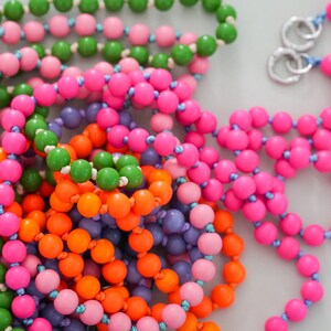 Könnte beinhalten: Nahaufnahme einer bunten Perlenkette mit grünen, rosa, orangefarbenen und violetten Perlen. Die Kette besteht aus mehreren Perlensträngen und ist verknotet.