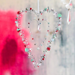 Pode incluir: Um quadro de arame prateado em forma de coração com um quadro de arame menor em forma de coração dentro. Os quadros são decorados com contas e cristais vermelhos, verdes e brancos.