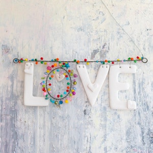 Può includere: Lettere in ceramica bianca che compongono la parola "LOVE" infilate su perline colorate e un filo. Le lettere sono appese su uno sfondo bianco testurizzato.