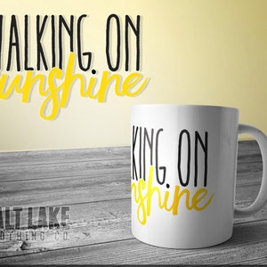 May include: White ceramic mug with black text that reads "Walking on Sunshine" in a yellow script font. The mug is sitting on a wooden surface.