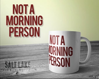 Może przedstawiać: Biały ceramiczny kubek z napisem "Not a Morning Person" w czerwonych literach. Kubek stoi na drewnianej powierzchni.