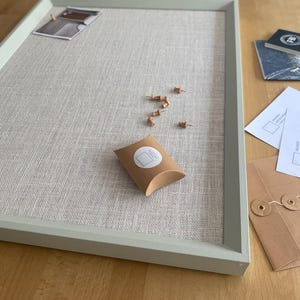 May include: A rectangular notice board with a light grey wooden frame and a textured beige linen fabric surface. The board includes a small brown cardboard gift box with a circular sticker, several wooden push pins, and a small photograph.