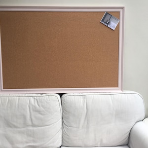May include: A large corkboard with a pale pink frame. A small photo is pinned to the upper right corner. The corkboard is resting on a white sofa. A small white table with a potted plant is to the right.