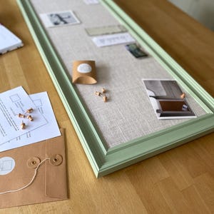 May include: A long, rectangular notice board with a light green wooden frame and a beige linen-textured fabric centre. The board features several small photographs, a brown paper gift box, and wooden push pins attached to the surface.
