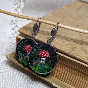 May include: A pair of black and silver earrings with a mushroom and green foliage design embroidered on a round black fabric base.