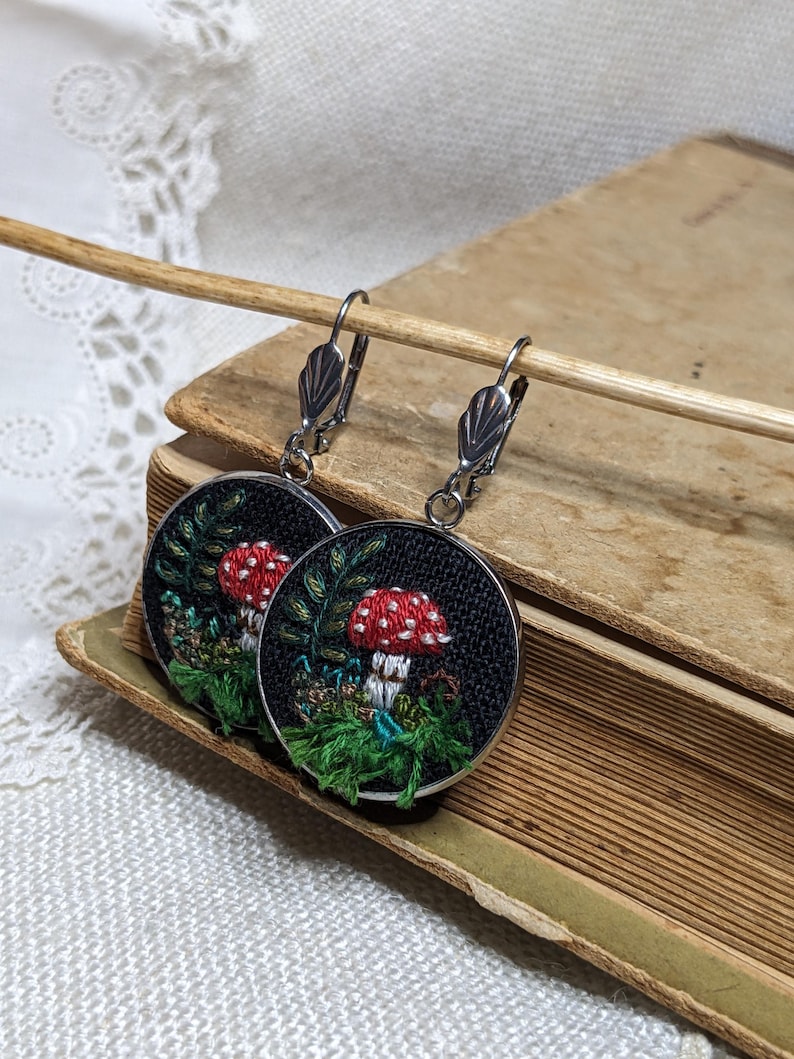 May include: A pair of black and silver earrings with a mushroom and green foliage design embroidered on a round black fabric base.