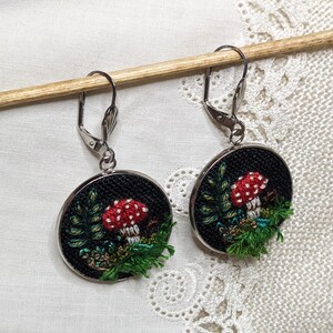 May include: A pair of silver dangle earrings with black circular pendants featuring embroidered red and white mushrooms with green foliage on a black background.