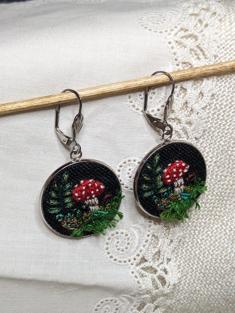 May include: A pair of silver dangle earrings with black circular pendants featuring embroidered red and white mushrooms with green foliage on a black background.