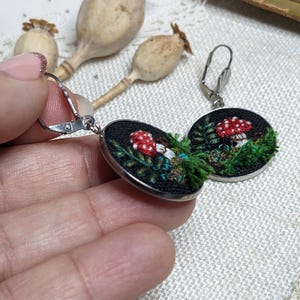 May include: A pair of silver hoop earrings with a black fabric circle featuring an embroidered mushroom and green foliage.