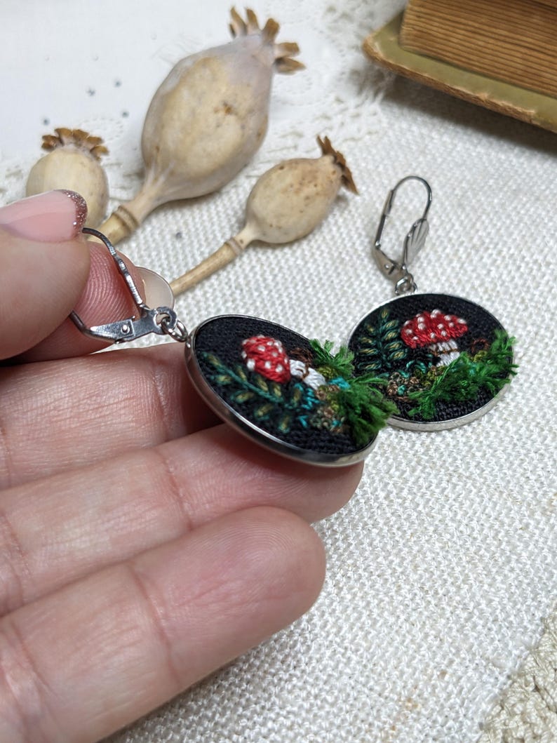 May include: A pair of silver hoop earrings with a black fabric circle featuring an embroidered mushroom and green foliage.