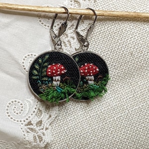 May include: A pair of silver hoop earrings with a black background and a red and white mushroom embroidered on each hoop.