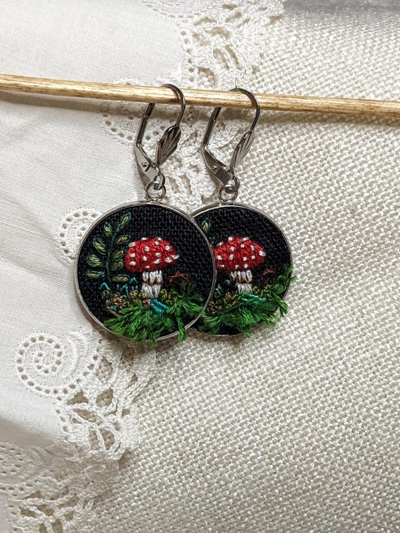 May include: A pair of silver hoop earrings with a black background and a red and white mushroom embroidered on each hoop.