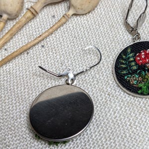 May include: A pair of silver earrings with a round, black background and a green and red embroidered mushroom design.