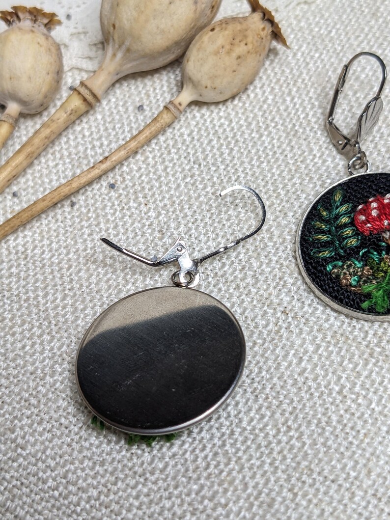 May include: A pair of silver earrings with a round, black background and a green and red embroidered mushroom design.