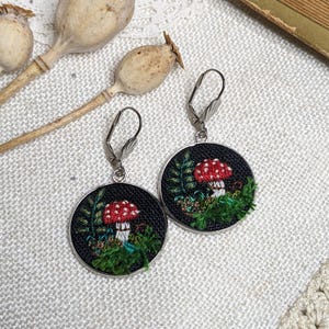May include: A pair of silver hoop earrings with black fabric circles featuring embroidered red and white mushrooms with green foliage.