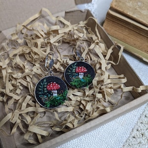 May include: A pair of earrings with a mushroom design. The earrings are made of silver and have a black background with green leaves and a red mushroom.