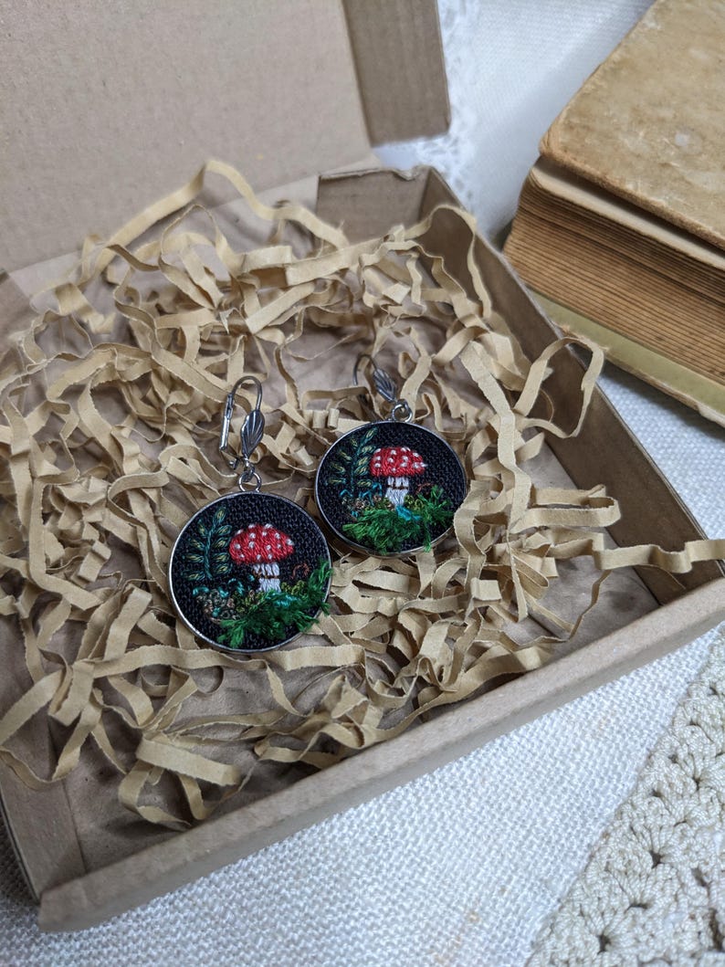 May include: A pair of earrings with a mushroom design. The earrings are made of silver and have a black background with green leaves and a red mushroom.