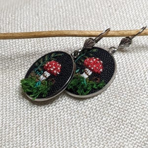 May include: A pair of silver earrings with a black background and embroidered red and white mushrooms with green foliage.