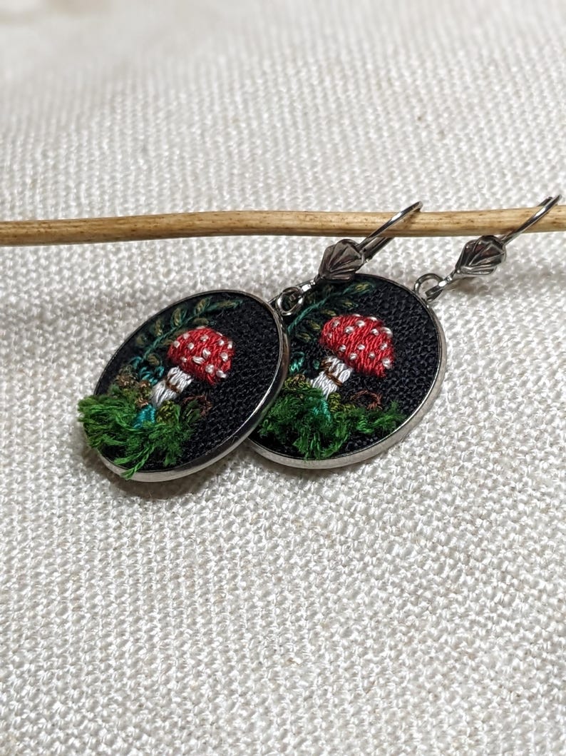 May include: A pair of silver earrings with a black background and embroidered red and white mushrooms with green foliage.