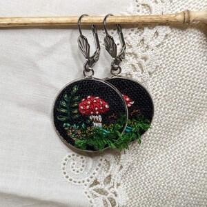 May include: A pair of silver hoop earrings with a black fabric circle featuring an embroidered red and white mushroom with green foliage.