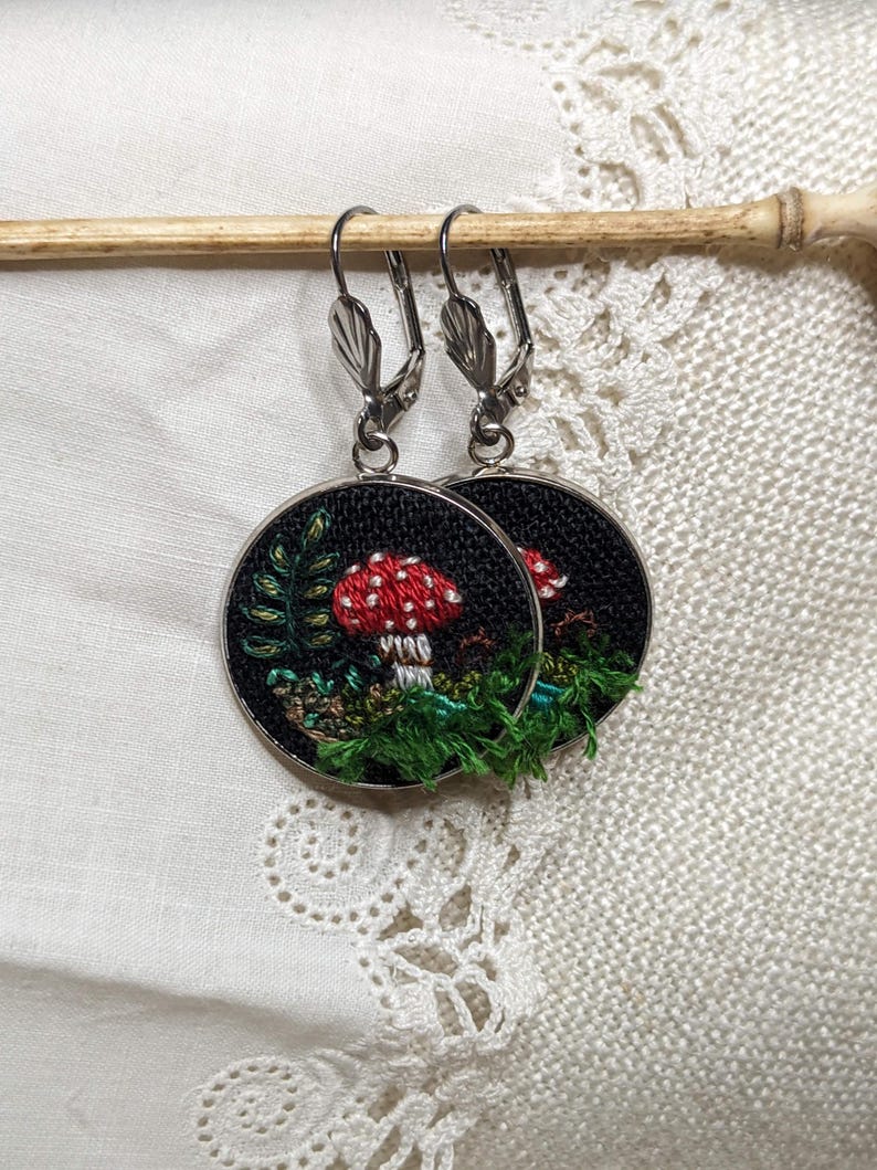 May include: A pair of silver hoop earrings with a black fabric circle featuring an embroidered red and white mushroom with green foliage.