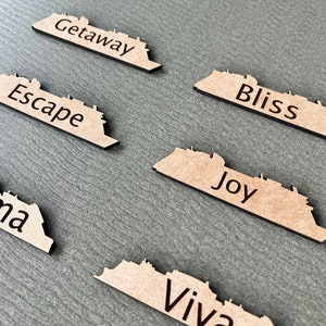 May include: Wooden cruise ship-shaped cutouts with black text. The cutouts are tan with black edges and feature words like "Getaway," "Escape," and "Bliss." They are arranged on a gray surface.