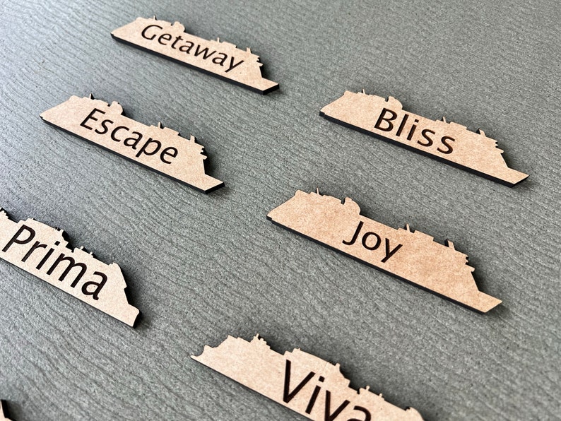 May include: Wooden cruise ship-shaped cutouts with black text. The cutouts are tan with black edges and feature words like "Getaway," "Escape," and "Bliss." They are arranged on a gray surface.