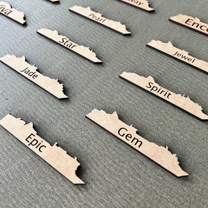 May include: A collection of laser-cut wooden name tags, each shaped like a stylized map. The tags are light brown with black text, featuring names like "Epic," "Gem," and "Spirit." They are arranged on a gray surface.
