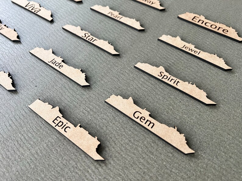 May include: A collection of laser-cut wooden name tags, each shaped like a stylized map. The tags are light brown with black text, featuring names like "Epic," "Gem," and "Spirit." They are arranged on a gray surface.