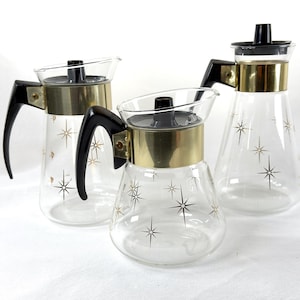 May include: Three vintage glass carafes with black handles, lids, and starburst designs. Each carafe has a gold-colored band around the middle. The carafes are clear, allowing the contents to be visible.