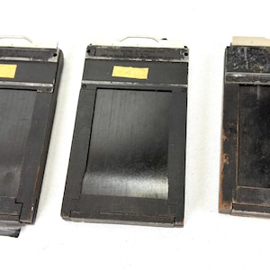 May include: Three vintage black wooden film holders with metal handles and small yellow labels. The holders have a rectangular design with a sliding mechanism to hold film. The wood shows signs of age and wear, suggesting they are antique.