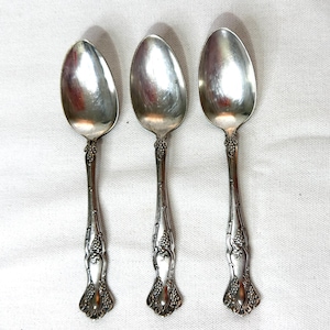 May include: Three silver spoons with ornate handles featuring a grape cluster design. The spoons have oval bowls and are arranged vertically against a white background. The spoons are likely vintage or antique.