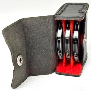 May include: A black leather case with three circular camera filters. The filters are black with white text that reads "COATED" and numbers 1, 2, and 4. The interior of the case is red.