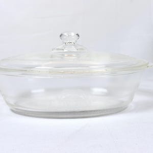 May include: A clear glass casserole dish with a matching lid. The dish is oval-shaped with small handles on either side. The lid has a small, round knob for easy handling. The dish is designed for cooking and serving.