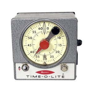 May include: A gray, square-shaped Time-O-Lite Master timer with a textured metal finish. The circular face features a cream-colored dial with black markings from 0 to 60 seconds, a red center pointer, a black adjustment knob, and two small metal switches.