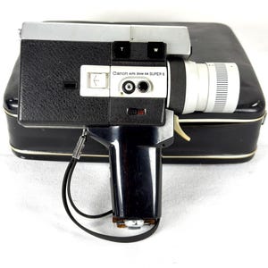 May include: A vintage Canon Auto Zoom 518 Super 8 camera with a black and silver body, a white lens, and a black handle. The camera is resting on a black case with a white trim. The camera has the text "Canon Auto Zoom 518 SUPER 8".