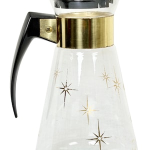 May include: A clear glass carafe featuring a gold-toned metallic band around the neck and a black plastic handle and lid. The glass body is decorated with a repeating pattern of gold-colored starburst designs scattered across the exterior surface.