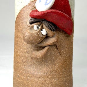 May include: A handmade ceramic mug with a sculpted face design. The mug features a red cap, a white and blue emblem with "FD" on it, and a textured brown surface. The rim is white with a blue trim. The mug is a unique piece of art.