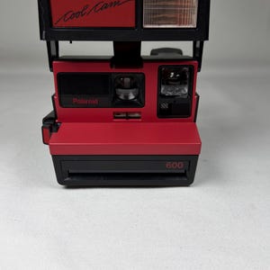 May include: A red and black Polaroid 600 Cool Cam instant camera. The camera has a built-in flash and the words "Cool Cam" and "Polaroid" are visible. The camera is designed for instant photography.