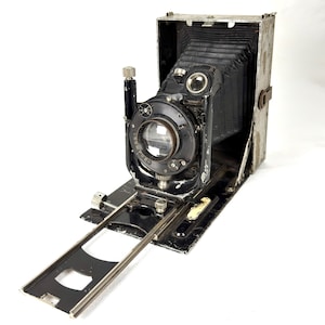 May include: An antique black and silver folding camera with a bellows design. The camera has a large lens and various dials and knobs. The camera is open, revealing the lens and internal components. The camera is on a black base with a silver rail.
