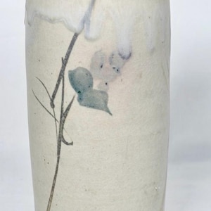 May include: A cylindrical ceramic vase with a cream-colored body and a light blue rim and base. The vase features a delicate floral design in shades of blue and gray, adding a touch of nature-inspired artistry.