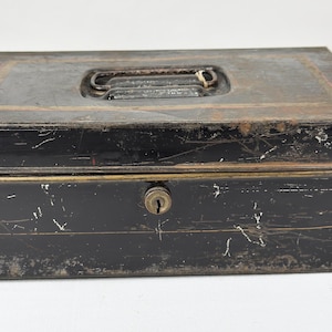 May include: A vintage black metal cash box with a handle and a keyhole. The box has a rectangular shape with a slightly rusted appearance, and a gold-colored trim around the top. The handle is made of metal and is located on the top of the box.