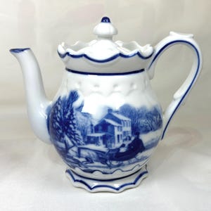 May include: White porcelain teapot with a blue winter scene design. The teapot features a scalloped rim and handle, and a matching lid with a small finial. The winter scene depicts a house, trees, and a horse-drawn sleigh.